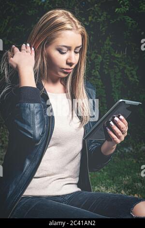 Blond haired studen doing e-learning in the park via her tablet PC. Vintage looking matte style image. Stock Photo
