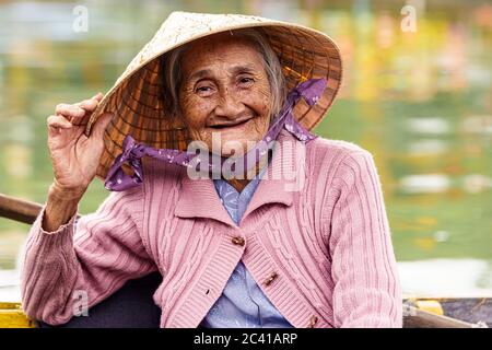 Old Vietnamese woman in violet sweater holds her hat while smiling Stock Photo