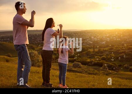 Affectionate family having fun blowing soap bubbles on mountain top at sunset. Parents and child playing together Stock Photo