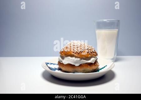 A traditional Scandinavian shrovetide bun with whipped cream, pearl sugar on top and a glass of milk on white table. Stock Photo