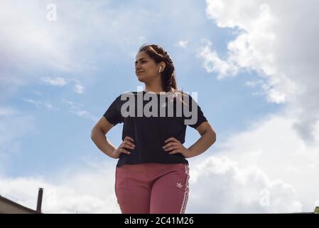 portrait of an Indian girl athlete, view from below, listens to music on headphones, against a background of clouds, and buildings Stock Photo