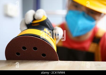 Masked carpenter grinds wood with sander machine Stock Photo