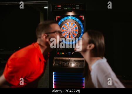 A beautiful young caucasian woman and a man lean over for a kiss and between them is a darts board. Love and recreation Stock Photo