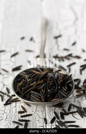 Heap of uncooked, raw, black wild rice grains in metal spoon on white rustic table background, selective focus Stock Photo