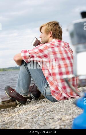 Young man sits by the water and drinks coffee Stock Photo