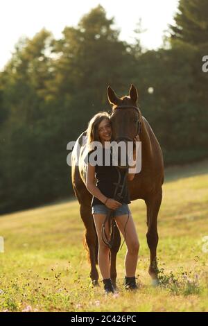 Smilling young girl with chestnut horse on meadow during summer sunset. Romanic scene Stock Photo