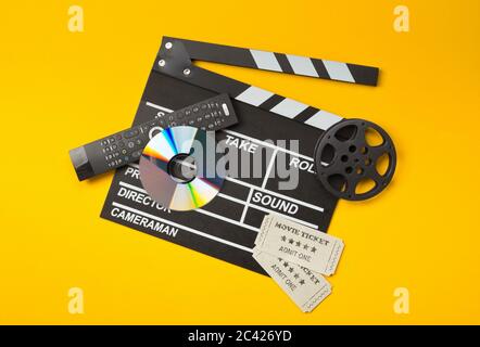 Single, black, open movie clapper or clapper-board with dvd movie disc, film reel, remote control and movie theatre tickets on yellow or orange backgr Stock Photo
