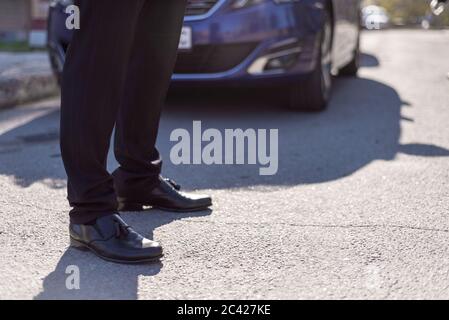 male legs in black leather shoes and black trousers, against the background of a silhouette of a car in blur Stock Photo