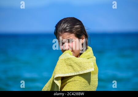 Portrait of preteen brunette girl, wet and wrapped in towel after swimming, against blue sea and sky bokeh background