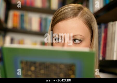 Young blonde woman in the middle of bookshelves peeks out from behind a book - search - library Stock Photo