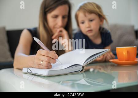 Young student mother studying from home, making notes in notebook  with her toddler daughter next to her. Stock Photo