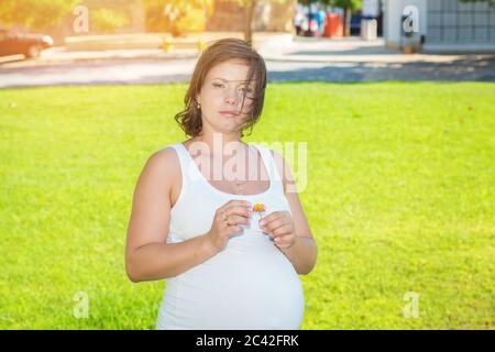 It is a boy or a girl. Pregnant Woman holding flower ripping its petals guessing the gender of her baby standing outdoors on a meadow in a park on sun Stock Photo