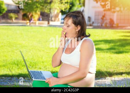 Tired young pregnant woman yawning while working on laptop sitting outdoors on a green grass meadow in a city park on a sunny day. Exhausted worker mo Stock Photo