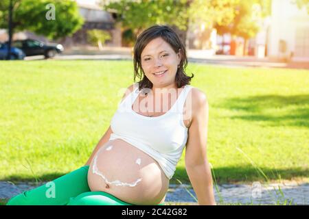 Belly of pregnant happy woman  with cream in shape of face with smile, woman applying moisturizing cream for stretch marks while sitting in park on gr Stock Photo