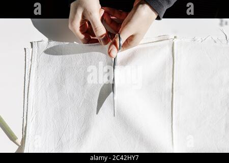 Top view of tailor working at table cutting a piece of cloth with scissors for making homemade protective masks Stock Photo