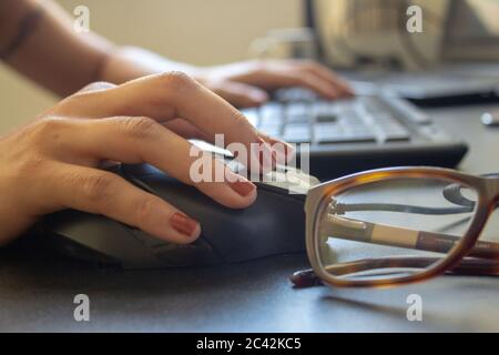 Closeup on the hand of a woman using her computer and glasses, with a keyboard, laptop and monitor on the background Stock Photo