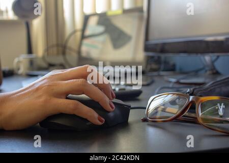Closeup on the hand of a woman using her computer and glasses, with a keyboard, laptop and monitor on the background Stock Photo