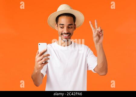 Happy cute african-american guy on vacation taking selfie to post online, holding smartphone, smiling and making peace sign at mobile camera, standing Stock Photo