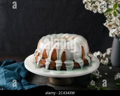Bundt cake with green matcha glaze on white marble cake stand over dark table with bouquet of blossoming cherry or pear branches. Copy space top over Stock Photo