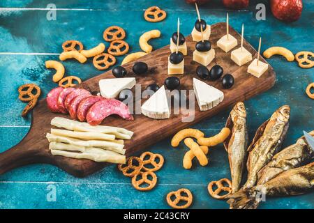 Sausage slices with cheese cubes, olives and crackers on a wooden board with dry fish on the blue table Stock Photo