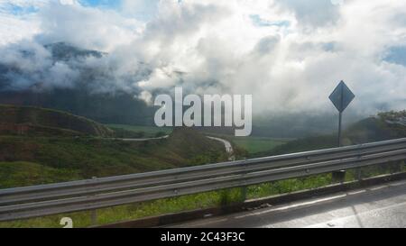 Cloudy landscape dresses from the edge of a road. Sunlight passing through the clouds over the green mountains with a road going down the mountain Stock Photo