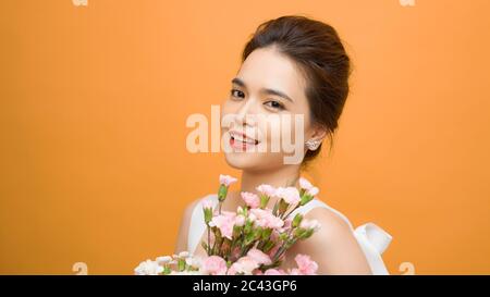 Beautiful woman in the white dress with flowers carnation in hands on a yellow background. She has gentle smile. Stock Photo