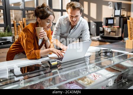 Two young managers or shop owners having some discussion working on a laptop at the counter of the shop or cafe. Small business management concept Stock Photo