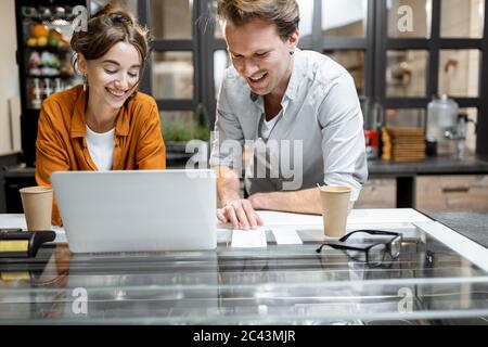 Two young managers or shop owners having some discussion working on a laptop at the counter of the shop or cafe. Small business management concept Stock Photo