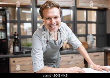 Portrait of a cheerful salesman in apron standing behind the counter of a small shop or cafe. Concept of a small business and work in the field of services Stock Photo