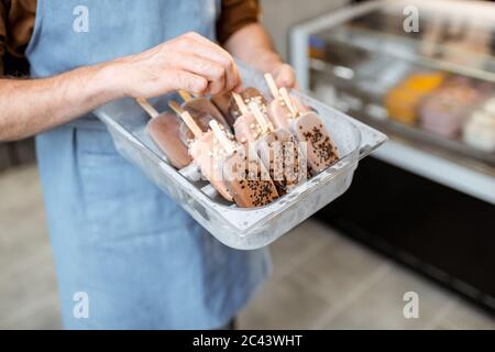 Seller holding a tray with chocolate ice cream on a stick in the shop, close-up on ice cream on a blue apron background Stock Photo