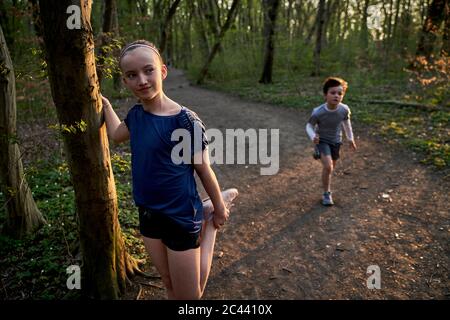 Girl looking away while stretching her leg by tree trunk against brother running in forest Stock Photo