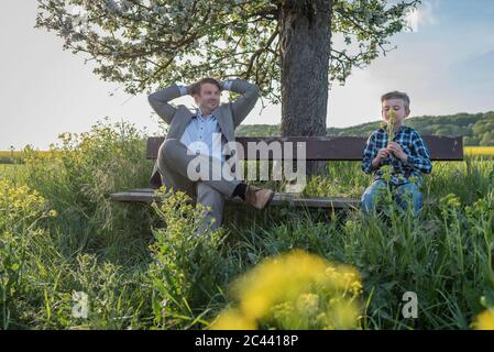 Relaxed man looking away while sitting by son smelling flowers on bench at park against sky Stock Photo