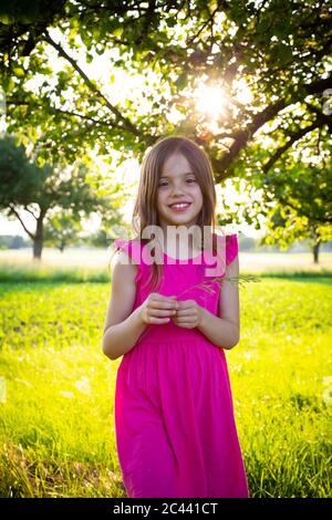 Portrait of little girl wearing vibrant pink dress smiling at camera with blade of grass in hands Stock Photo