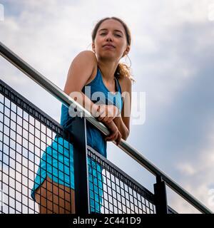 Portrait of young attractive woman wearing tank top and shorts leaning on railing against sky Stock Photo