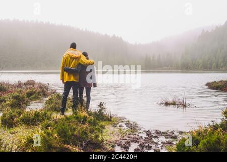 Full length rear view of young couple standing while looking at river near forest during rainy season Stock Photo