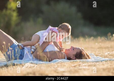 Happy woman carrying baby girl while lying on blanket on meadow during sunny day