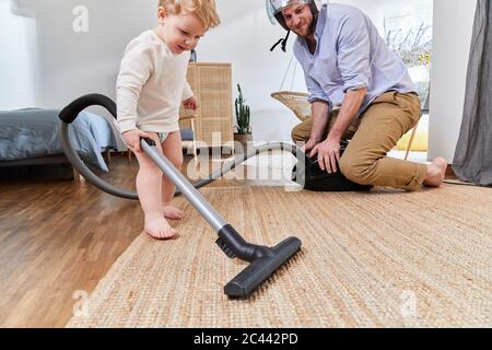 Man looking at cute toddler son cleaning carpet with vacuum cleaner in living room at home Stock Photo