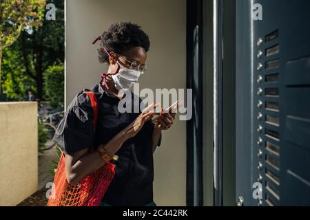 Young woman wearing textile protective mask standing in front of entry door using smartphone Stock Photo