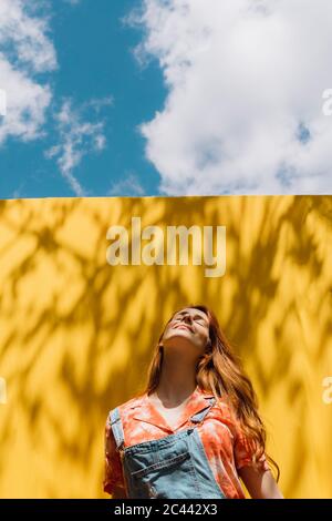 Relaxed young woman standing over yellow wall against cloudy sky Stock Photo