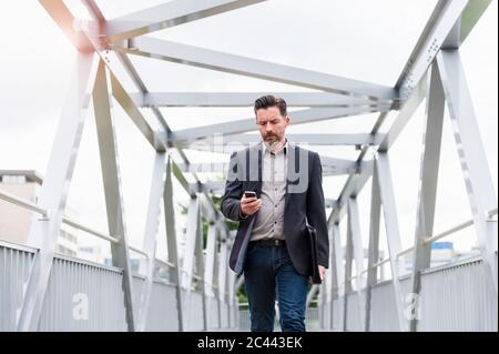 Confident handsome businessman using smart phone while walking on footbridge in city Stock Photo