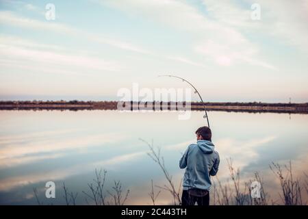 Rear view of teenage boy wearing hooded shirt fishing with rod in lake against cloudy sky Stock Photo