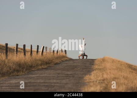 Mid distance view of mature man doing headstand on road against clear sky Stock Photo