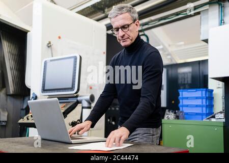 Male supervisor reading document while using laptop in factory Stock Photo