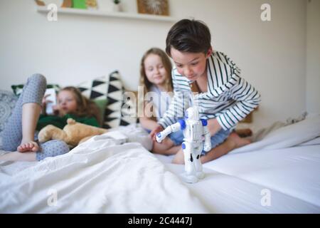 Excited little boy crouching on bed playing with toy robot while his sisters relaxing in the background Stock Photo