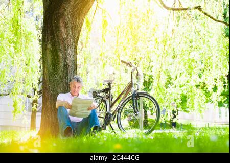 Senior man leaning against tree trunk in a park reading newspaper Stock Photo