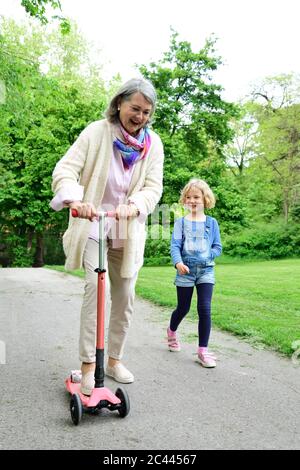 Cheerful senior woman riding push scooter while granddaughter walking in park Stock Photo