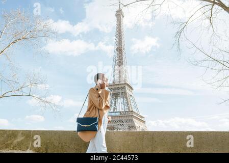 Young woman using smart phone while walking on bridge with Eiffel Tower in background, Paris, France Stock Photo