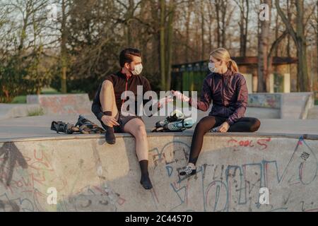 Woman giving man hand sanitizer while sitting at skateboard park during COVID-19 Stock Photo