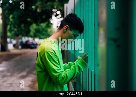 Thoughtful young man looking down while standing by metallic fence in city Stock Photo
