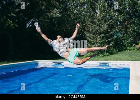 Shocked young man holding drink falling in swimming pool against trees Stock Photo